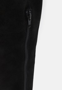 Detailní záběr na černou suede látku s vertikálním černým zipem a hladkým švem na bílém pozadí.