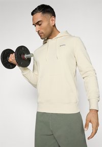 Haut à capuche crème avec cordons de serrage et poche kangourou, associé à un pantalon de survêtement vert. Personne tenant un haltère noir.