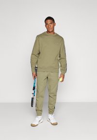 Conjunto de sudadera y pantalones jogger en verde oliva, con un corte relajado, puños acanalados y detalles de franjas laterales. Accesorios con una raqueta de tenis y una pelota.