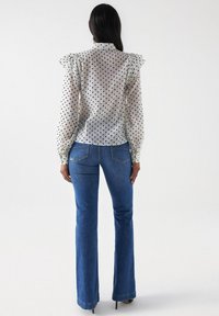 Blusa blanca transparente con lunares negros, hombros con volantes, mangas largas, combinada con jeans acampanados azules, con bolsillos traseros visibles y dobladillo.