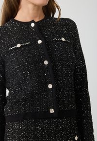 Čierna pletená bunda s flitrovou textúrou, striebornými gombíkmi a vlnitými detailmi na vreckách. Žebrovaný lem a manžety.
