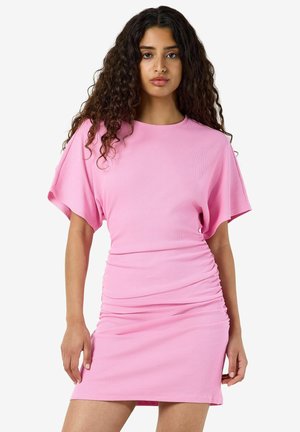 Femme aux longs cheveux bouclés foncés portant une robe courte rose avec des manches larges et des détails froncés sur la jupe.