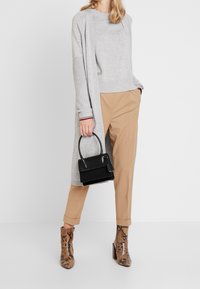 Woman wearing gray long cardigan, beige pants, snake-print ankle boots, holding black handbag against white background.