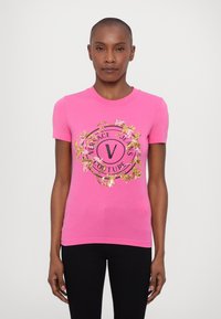 Rozā īsām piedurknēm t-krekls ar centrālu apļveida grafiku, uz kura redzams "Versace Jeans Couture" un ziedu akcenti zeltā un melnā krāsā.