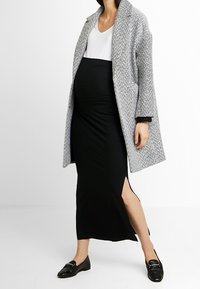 Femme enceinte portant un haut blanc, une jupe noire taille haute avec une fente sur le côté, un manteau gris à motifs et des mocassins noirs sur fond blanc.
