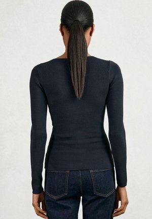 Kvinna med långt mörkt hår i hästsvans, iförd en figurnära svart långärmad topp och mörkblå jeans, som står mot en enkel bakgrund.