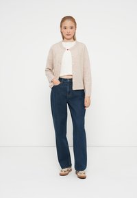 Beżowy sweter z dzianiny z okrągłymi guzikami, noszony na białym topie crop; zestawiony z ciemnoniebieskimi dżinsami o wysokim stanie i beżowymi loafersami.