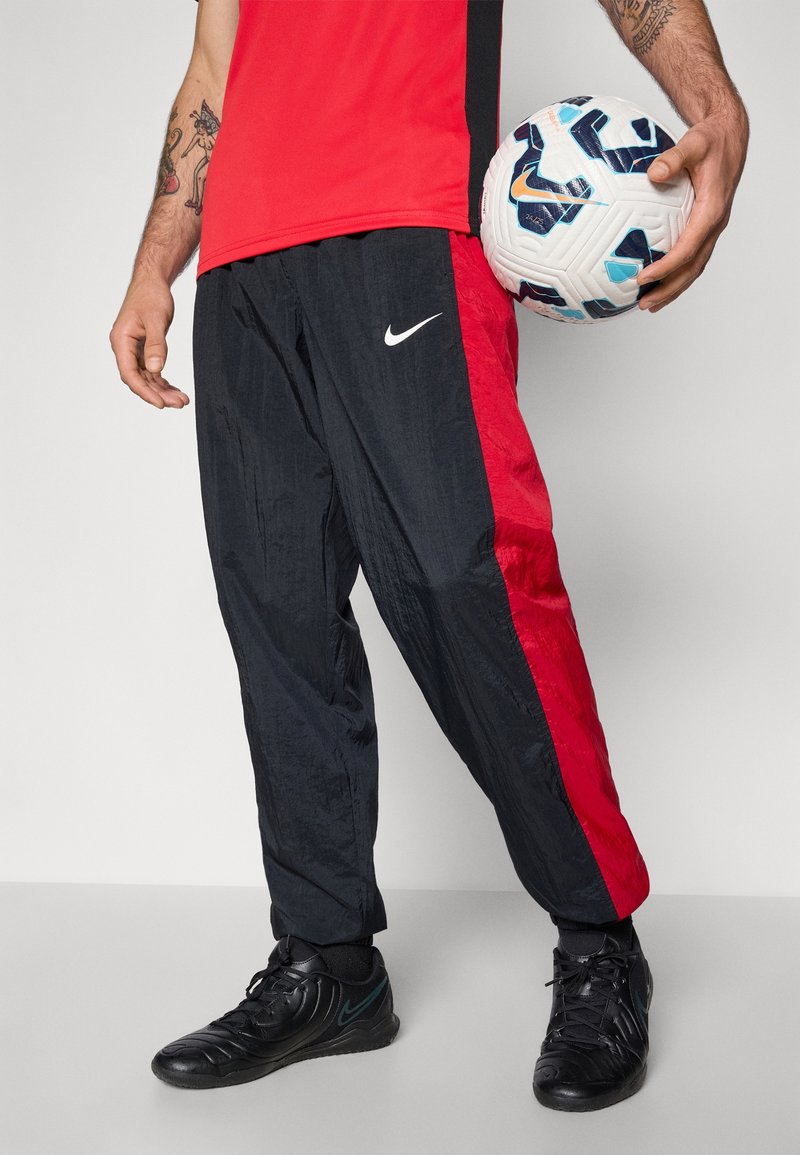Homme avec des tatouages tenant un ballon de football blanc et bleu, portant un pantalon de sport rouge et noir, un t-shirt rouge et des baskets noires.