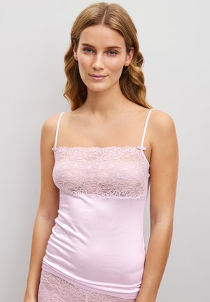 Jeune femme aux cheveux châtain clair portant un caraco rose bordé de dentelle et un bas assorti, debout devant un fond gris uni.