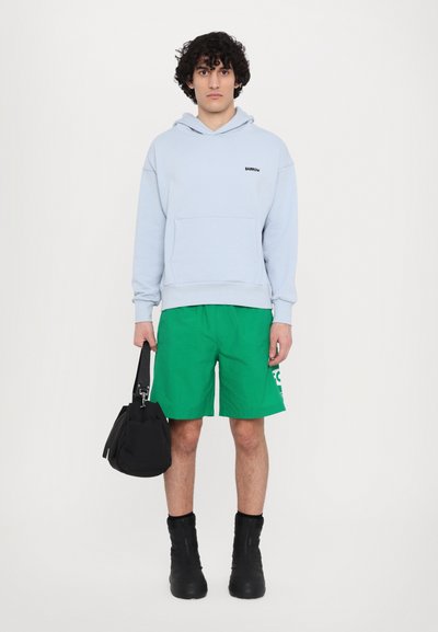 Sudadera azul clara con bolsillo frontal y logo negro, pantalones cortos verdes, mochila negra y botas altas negras. Diseño de atuendo sencillo y casual.