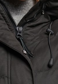 Primer plano de la cremallera de una chaqueta de invierno negra, botón a presión y cordón ajustable con regulador, llevada sobre un suéter gris de punto.