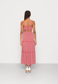 Röd blommig maxiklänning med tunna axelband, lager-på-lager-design och öppen ryggdetalj. Lättviktigt tyg, intrikat mönster och flödande silhuett.