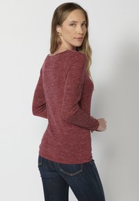 Burgundy színű, hosszú ujjú felső, puha, strukturált anyagból készült, kerek nyakkivágással, sötét farmernadrággal párosítva, kiemelve az alakot.