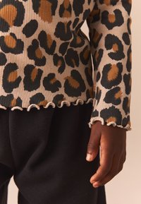 Leopard-patterned ribbed top in beige, featuring brown and black spots, with a ruffled hem and black cotton pants, hand resting beside.