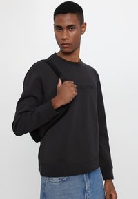 Black sweatshirt with a crew neckline, featuring embossed branding. Model holds a black strap backpack. Light blue denim jeans visible.