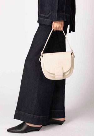 Beige leren crossbodytas met een flapsluiting, voorzien van een textuureffect en een klein riempje, vastgehouden door een persoon in donker denim met wijde pijpen.