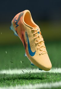 Nike-Fußballschuh in leuchtendem Gelb mit blauen Akzenten, strukturiertem Obermaterial und Stollen, hoch über Gras mit Rasenresten schwebend.