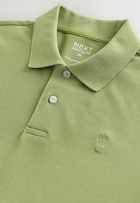 Vaaleanvihreä poolopaita, joka on valmistettu teksturoidusta kankaasta. Paidassa on kaulus, kaksi nappia ja vasemmalla puolella kirjailtu kuvio.