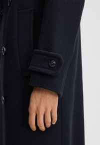 Marineblauwe wollen jas met een geknoopte manchet, waarbij een zijaanzicht van de mouw en een hand te zien is. De jas heeft een gladde, gestructureerde stof en twee zijzakken.