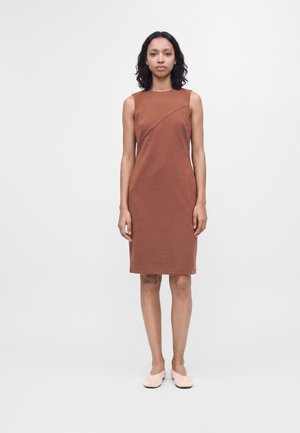 Femme debout, droite, vêtue d'une robe marron sans manches jusqu'aux genoux et de ballerines beige clair sur un fond clair uni.
