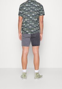 La camisa de manga corta presenta un patrón geométrico en verde y gris; combinada con unos pantalones cortos de mezclilla gris bicolor y zapatillas deportivas altas verdes.