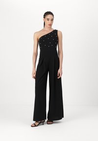 One-shoulder black jumpsuit with pearl embellishments, wide legs, and pleated design. Worn with black strappy heels, smooth texture.