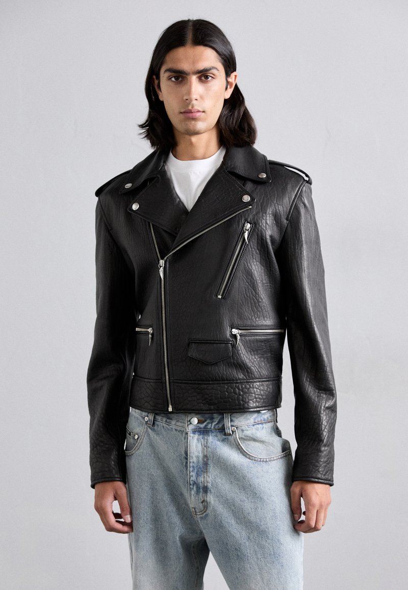 Black leather motorcycle jacket featuring textured material, silver zippers, and an asymmetrical design. Paired with light blue denim jeans.