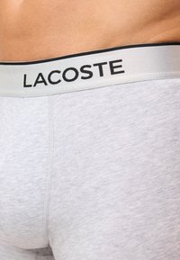 Grå bomullsboxershorts med myk tekstur og elastisk midje med "LACOSTE" trykt i svart på en hvit bakgrunn.