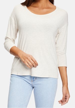 Kvinde iført en lysebeige langærmet top med rund hals og lyseblå jeans, vist fra skuldre til hofter.