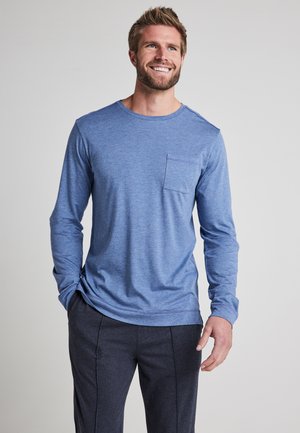 Long-sleeve blue cotton shirt with a crew neckline and a chest pocket. Smooth texture with a relaxed fit design, paired with dark trousers.