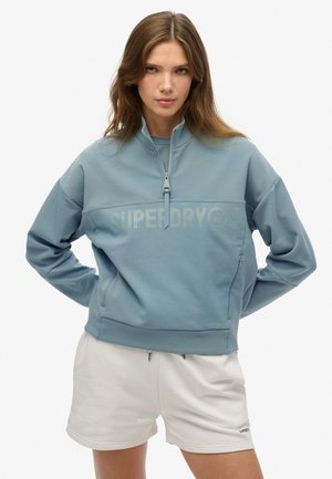 Felpa zip blu chiaro con colletto alto, logo frontale, maniche lunghe e vestibilità rilassata. Abbinata a pantaloni corti bianchi.