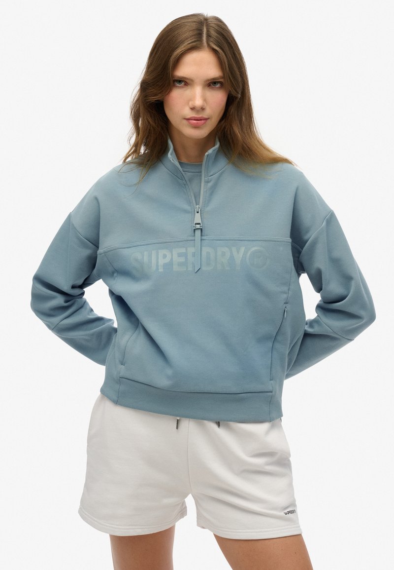 Lichtblauwe rits sweatshirt met hoge kraag, voorlogo, lange mouwen en een ruime pasvorm. Gecombineerd met witte shorts.