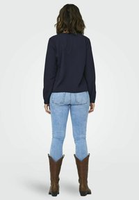 Haut à manches longues bleu marine, jean skinny bleu clair et bottes de cow-boy marron. L'ensemble est vu de l'arrière, mettant en valeur la coupe.