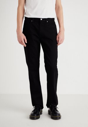 Oseba nosi črne ravne hlače, belo majico z zavihkom in črne vezalke s rumenimi šivi na navadni beli podlagi.