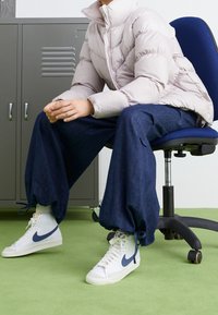 Luftig, ljusrosa vadderad jacka med ett mönstrat tyg, kombinerad med vida, mörkblå denimbyxor och vita sneakers med en marinblå swoosh.