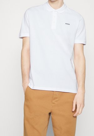 Man wearing white short-sleeve polo shirt with "HUGO" logo and mustard-colored pants, hands casually in pockets, on light background.