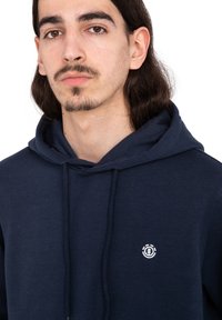 Sudadera azul marino hecha de tela suave, con capucha ajustable y un pequeño logotipo blanco en el pecho. Diseño simple con un corte holgado.
