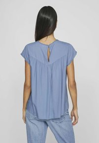 Blusa azzurro chiaro con maniche a cappuccio, dettaglio di apertura a chiave sul retro e bordo testurizzato attorno al collo. Tessuto morbido con vestibilità ampia.