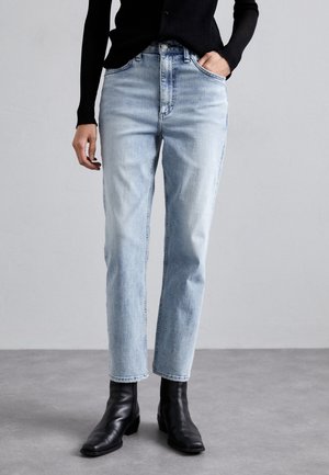 Personne portant un jean droit bleu clair, un haut noir à manches longues et des bottines noires, debout sur un sol gris devant un mur uni.