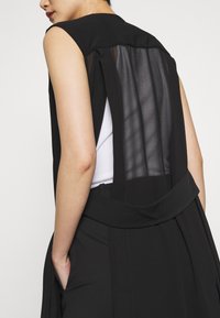 Svart ärmlös topp med en genomskinlig bakpanel, med en strukturerad krage, och dekorerad med en bred midjeband och sidofickor.