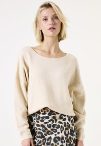 Pull beige avec un décolleté large et des manches longues ; texture douce. Assorti à une jupe en imprimé léopard texturé avec des accents foncés et dorés.