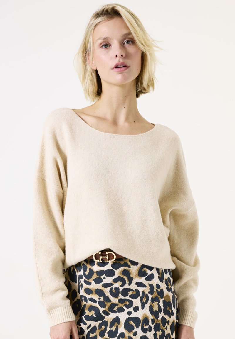 Beiger Pullover mit weitem Halsausschnitt und langen Ärmeln; weiche Textur. Kombiniert mit einem strukturierten Leopardenmuster-Rock mit dunklen und goldenen Akzenten.