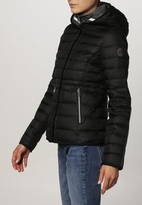Sort pufferjakke med hætte, der har horisontale quiltede sektioner, lynlåslommer på siden og et logo-patch på ærmet.