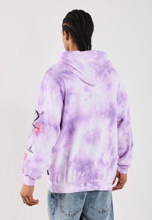 Osoba z warkoczami, ubrana w fioletową bluzę z kapturem tie-dye z różowym kwiatowym haftem na rękawach oraz jasnoniebieskie jeansy, stoi tyłem.