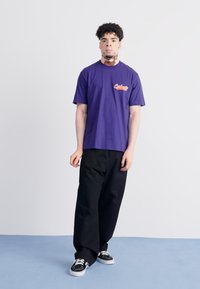 Purple cotton t-shirt with a rounded neckline and a small red logo on the left side, paired with loose-fitting black pants and black sneakers.
