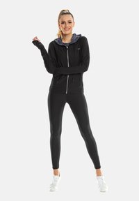 Zwarte sportjas met een grijze capuchon, ritssluiting aan de voorkant en duimgaten, gecombineerd met zwarte leggings en witte sneakers, geschikt voor sportieve activiteiten.