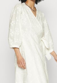 Bílá zavinovací šaty s 3/4 nabíranými rukávy, květinovou výšivkou, výstřihem do V a detailem na zavázání v pase. Vyrobeno z lehké tkaniny. Viditelný zlatý řetízek na krk.