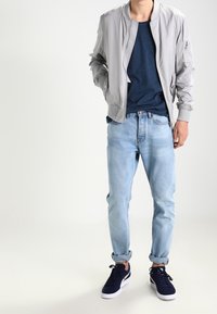 Jasnoszara kurtka bomber, niebieska koszulka, jasnoniebieskie dżinsy z podwiązanym nogawkami i granatowe sneakersy. Gładki materiał, luźny krój i swobodny styl.