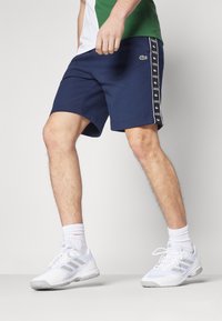 Man wearing navy Lacoste shorts and white Adidas sneakers standing against a plain white background.