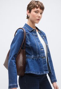 Μια εφαρμοστή, μπλε τζιν ζακέτα με σχεδιασμό πεπλούμ, ασημένια κουμπιά και καφέ πλεκτή τσάντα, αποτυπωμένα πάνω σε ένα μονόχρωμο φόντο.